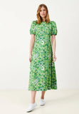 A woman in a green patterned Jumperfabriken Dress Philomena by BUTIK KUBIK, made from a cotton blend with puffed shoulders, and white sneakers, stands against a white background.