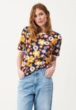 A woman with shoulder-length light brown hair wears the Jumperfabriken Edith Top Navy by BUTIK KUBIK, featuring a bold yellow, orange, and white retro pattern, paired with light blue jeans. She stands hands-in-pockets against a plain background.