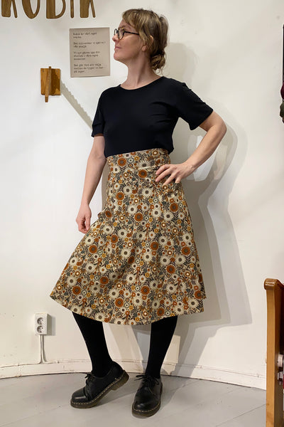 Indoors, an individual models a 50s-inspired Kubik skirt Twist Gerbera from BUTIK KUBIK, showcasing its brown, white, and orange floral design. They pair it with a black top, tights, and shoes. The setting includes a simple wall with a small sign and shelf in the background.