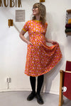 A person poses indoors, wearing the Kubik dress Twist Popsicle by BUTIK KUBIK—an orange dress with a pink circle pattern made from GOTS-certified fabric. Paired with black tights and shoes, they hold out part of the dress. A small shelf with folded items is partially visible on the right.