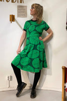 Wearing a Kubik dress Twist the leaf from BUTIK KUBIK, featuring a 50s-inspired green design with large leaf patterns made from GOTS-certified fabric, a person stands in black tights and shoes against a white wall. Their shoulder-length wavy hair adds to their stylish look as they gaze to the side.