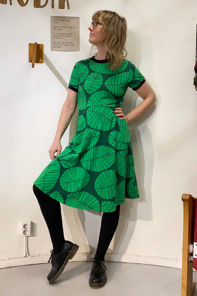 Wearing a Kubik dress Twist the leaf from BUTIK KUBIK, featuring a 50s-inspired green design with large leaf patterns made from GOTS-certified fabric, a person stands in black tights and shoes against a white wall. Their shoulder-length wavy hair adds to their stylish look as they gaze to the side.