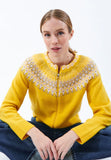 A person sits cross-legged, wearing the Jumperfabriken Audrey Cardigan Yellow by BUTIK KUBIK—an exquisitely knit sweater made from soft merino wool—with a detailed, patterned yoke in white, pink, and gray tones. The background is plain white.