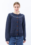 A woman wearing the BUTIK KUBIK Jumperfabriken Gullvor cardigan in blue, featuring a navy blue pattern and brown buttons, paired with blue jeans, stands against a plain white background, smiling gently at the camera.