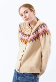 A woman wearing the BUTIK KUBIK Jumperfabriken Joelle cardigan in sand—a cream-colored knit with a pink, brown, and yellow geometric patterned yoke—paired with blue jeans and her hair parted in the middle, stands against a white background.