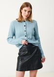 A woman is seen wearing the BUTIK KUBIK Jumperfabriken Luca cardigan in turquoise—a soft wool, button-up knit—styled effortlessly with a black leather skirt as she stands against a plain white background.