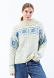 A woman wearing the BUTIK KUBIK Jumperfabriken Katie jumper—a cream-colored knit sweater with blue patterns—paired with blue jeans, standing against a plain white background.