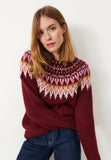 A woman with straight, shoulder-length blonde hair wears the Jumperfabriken Veda jumper in burgundy by BUTIK KUBIK, featuring a pink, white, and brown geometric klassiskt okmönster, paired with dark blue jeans and standing against a plain white background.