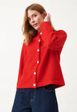 A woman wears the Jumperfabriken Cicely cardigan in red by BUTIK KUBIK— a bright, stickad kofta with white buttons crafted from soft merinoull— paired with dark blue jeans. She stands against a plain white background, one hand touching her hair.