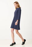 A woman wearing the BUTIK KUBIK Jumperfabriken Casey dress navy, featuring a patterned design with three-quarter sleeves, and black shoes walks to the right against a plain white background.