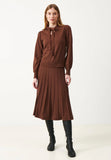 A woman stands against a plain white background, wearing a brown long-sleeve top with a bow at the collar, the Jumperfabriken Klara skirt in brown by BUTIK KUBIK, and black knee-high boots.