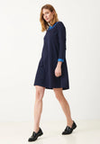 A woman with light brown hair wears the Jumperfabriken Tindra dress navy by BUTIK KUBIK, featuring a blue collar and cuffs, paired with black loafers as she stands against a plain white background.