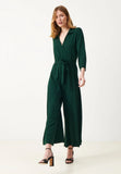 A woman stands against a plain white background wearing the Jumperfabriken Blakely jumpsuit green by BUTIK KUBIK—a dark green, long-sleeve wrap jumpsuit with a tie waist and wide legs—paired with black heeled sandals.
