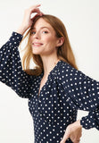 Woman smiling in the Jumperfabriken Cosima dress by BUTIK KUBIK—a navy blue midiklänning with white polka dots in ekologisk bomull—posing with her right hand in her hair against a plain, light background.
