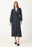 A woman stands against a plain backdrop wearing the Jumperfabriken Cosima dress by BUTIK KUBIK, a long-sleeved navy blue polka dot midi dress in organic cotton, paired with black flats.