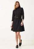 A woman wearing the Jumperfabriken Kim dress check by BUTIK KUBIK—a black, knee-length dress with a brown checkered pattern, high collar, three-quarter sleeves, and belted waist—paired with black loafers, stands against a plain white background.