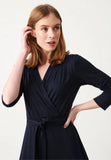 A woman with straight, shoulder-length light brown hair wears the Jumperfabriken Tamara dress navy from BUTIK KUBIK—a classic wrap dress with three-quarter sleeves and a waist tie. Looking slightly to the side, she touches her hair with one hand, accentuating the feminine silhouette of her outfit.