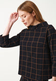 A woman with straight light brown hair wears the Jumperfabriken Tara jumper check black by BUTIK KUBIK, which features a brown graphic check pattern. With a relaxed silhouette, she faces left, gently touching her head and appearing thoughtful.