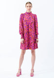 A woman stands against a plain white background, wearing the Jumperfabriken Julie dress in pink by BUTIK KUBIK—a vibrant pink and orange paisley-patterned dress featuring a high neckline and long sleeves—paired with matching pink platform heels.