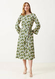 A woman stands against a plain white background wearing the Jumperfabriken Lenita dress green by BUTIK KUBIK—a floral-patterned, green, white, and yellow midiklänning featuring long sleeves and a waist tie. Crafted from ekologisk bomull, this mönstrad klänning combines style with sustainability. She completes the look with white heels.