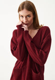 A woman with light brown hair faces the camera with a gentle, thoughtful expression, holding the sleeve of her Jumperfabriken Cordelia cardigan in burgundy by BUTIK KUBIK near her mouth. The cardigan is loose-fitting with two front pockets, and the background is plain white.
