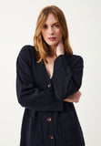 A woman with light brown hair wearing the Jumperfabriken Cordelia cardigan in navy from BUTIK KUBIK stands against a plain white background, looking at the camera with one hand touching her neck.