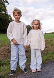 Two young children stand outdoors on a path, holding hands. Both wear cream-colored Jumperfabriken Bo jumpers from BUTIK KUBIK, crafted from soft Swedish wool and inspired by classic designs. The background features green grass, trees, and a cloudy sky.
