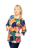 A woman with long blonde hair in a side braid smiles against a white background, wearing the BUTIK KUBIK Cissi och Selma top Vendela Skogsdröm with 3/4 sleeves and colorful shapes, paired with dark jeans.