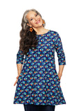 A smiling woman with long gray hair wears orange flower earrings and the BUTIK KUBIK Cissi och Selma Anita Midsommardans Blå dress, hands in pockets, tilting her head against a white background.