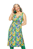 Smiling woman with gray hair wears the Cissi och Selma Anna Dress Illusion Färgglad by BUTIK KUBIK—a sleeveless, colorful floral dress—paired with green earrings and tights, standing against a white background.