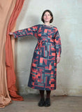 A woman wearing the Palava Dress Beryl Raspberry Highwire by BUTIK KUBIK stands indoors, holding back an orange curtain against a textured pale wall and worn wooden floor.