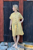 A barefoot person stands on rocks before a rustic wooden wall, wearing the oversized Kubik blouse dress block print yellow by BUTIK KUBIK, with buttons and pockets, looking to the side with one hand in a pocket.