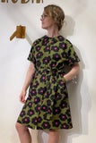 A person wearing glasses stands against a white wall, dressed in the BUTIK KUBIK Kubik blouse dress Black Flower with bold black and pink floral patterns.