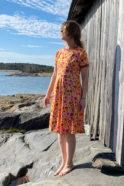 A barefoot woman in the Kubik dress Twist 1966 by BUTIK KUBIK stands on rocky ground near a weathered wooden building, her hair blowing in the wind as she faces a lake with trees and blue sky in the background.