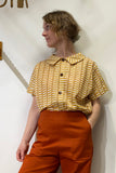 A person stands against a white wall, wearing the BUTIK KUBIK Kubik blouse block print yellow with a wide collar and brown buttons, paired with high-waisted orange pants. They look to the side with one hand in their pocket.