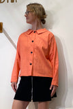 A person wearing glasses, the Kubik blouse linen peach—an oversized orange button-up long-sleeve blouse with a wide collar made from 100% linen by BUTIK KUBIK—and a black skirt with a front zipper stands against a white wall, looking to their left.