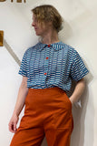 A person stands against a white wall, wearing the BUTIK KUBIK Kubik blouse block print petrol, an oversized cotton blouse with blue-and-white patterns and red buttons, paired with high-waisted orange pants. They look to the side with one hand in their pocket.