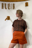 A woman with short blonde hair and glasses stands by a white wall with "KUBIK" above wooden hooks, wearing BUTIK KUBIK's oversized Kubik blouse floral viscose in black and red, styled with orange shorts and hands in her pockets, looking to the side.