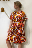 A woman in the Kubik blouse dress Groovy by BUTIK KUBIK stands against a white wall. The short-sleeved, knee-length dress features a bold swirl print, pockets, and is crafted from soft cotton satin as she gazes thoughtfully to the side.