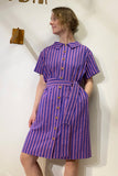 A person stands against a white wall wearing the BUTIK KUBIK Kubik blouse dress purple stripe with vertical purple and pink stripes, a matching belt, and rounded collar, looking to the side and smiling slightly.