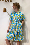 A woman stands against a white wall wearing the Kubik blouse dress vintage bluebell by BUTIK KUBIK, a short-sleeved, knee-length dress sewn in Sweden from blue, green, and white deadstock vintage fabric. She looks to the side with one hand in her pocket.