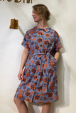 A woman stands against a white wall, wearing the Kubik blouse dress rusty flower by BUTIK KUBIK—an oversized, knee-length dress with short sleeves, a blue base with orange floral pattern, belted waist, and her hand in the pocket while looking to the side.