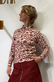 A woman in glasses stands sideways, wearing the BUTIK KUBIK Kubik sweater Botan Burgundy— a 60s-inspired knit top with a red and white botanical pattern—paired with a red skirt featuring a front zipper. Her left hand rests in her pocket as she looks to the left. The outfit is crafted from Oeko-Tex certified cotton and made in Sweden.