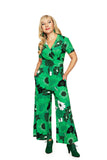 A woman with blonde loose curls wears the Cissi och Selma Britta jumpsuit 1960 Grön by BUTIK KUBIK, featuring a green 1960s floral print, V-neck, short sleeves, and wide legs. She pairs it with green heels against a white background, hands in pockets.