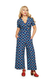 A woman with long blonde hair wears the Cissi och Selma Britta jumpsuit Midsommardans Blå by BUTIK KUBIK, styled with red earrings and red heeled shoes, standing with her hands in her pockets against a white background.