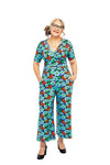 A woman with gray hair and glasses stands confidently in a vibrant BUTIK KUBIK Cissi och Selma Britta jumpsuit, featuring blue, red, and orange floral patterns and a stylish V-ringning. Smiling warmly with hands in pockets, she wears black pumps against a plain white background.