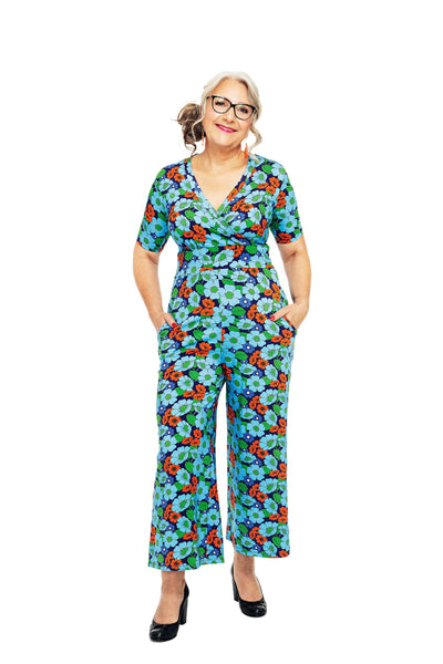 A woman with gray hair and glasses stands confidently in a vibrant BUTIK KUBIK Cissi och Selma Britta jumpsuit, featuring blue, red, and orange floral patterns and a stylish V-ringning. Smiling warmly with hands in pockets, she wears black pumps against a plain white background.