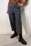 A person stands against a white wall wearing BUTIK KUBIK Kubik trousers wool in a gray check, a colorful floral long-sleeve top, black socks, and black lace-up shoes. Their hands are relaxed by their sides.