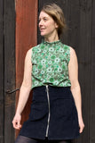A woman stands in front of a dark wooden wall, smiling and looking to her left. She wears the BUTIK KUBIK Kubik top cineast spring onion in green with a black skirt featuring a front zipper, one hand relaxed by her side.