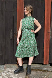 Wearing the BUTIK KUBIK Kubik skirt Twist Spring onion Midi, a person in black boots stands on stone steps before a rustic dark wooden door, looking to the side with one hand on their hip.